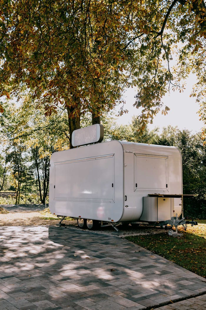 White mobile food trailer under trees in a sunny park, ideal for outdoor events or food festivals.
